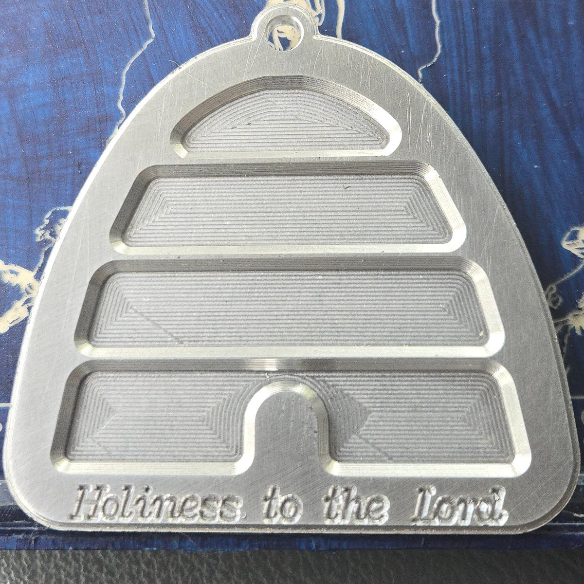 A silver metal beehive-shaped pendant with the inscription 'Holiness to the Lord' at the bottom, displayed on a blue backing card inside a clear plastic case.