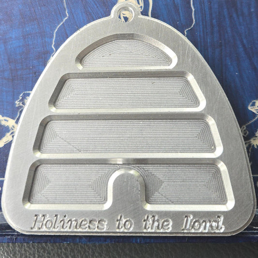 A silver metal beehive-shaped pendant with the inscription 'Holiness to the Lord' at the bottom, displayed on a blue backing card inside a clear plastic case.